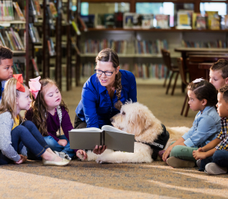Lehrerin mit Therapiehund liest einer Gruppe Kinder in der Bibliothek aus einem Buch vor